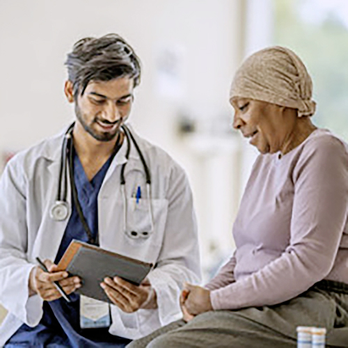 Oncologist Discussing Medications with a Senior Patient A male Oncologist of Middle Eastern decent sits with a senior patient as they discuss her medications. The patient is dressed casually and wearing a head scarf to keep her warm as she looks at the tablet in the doctors hands, and her prescriptions can be seen sitting out on the exam table beside her.